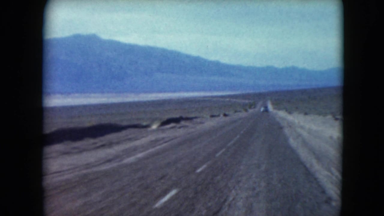 1961 Nevada 8mm road-trip view of a straight desert highway leading toward blue mountain ridges under a pale sky.