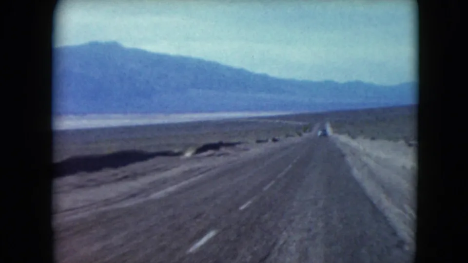 1961 Nevada 8mm road-trip view of a straight desert highway leading toward blue mountain ridges under a pale sky.