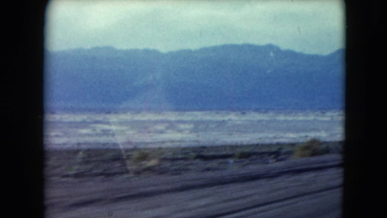 Moving 8mm view across Nevada desert flats, with scrub near the road and a blue mountain range in the distance.