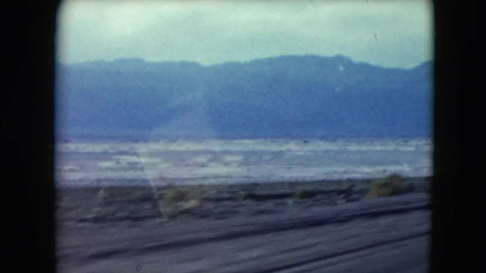 Moving 8mm view across Nevada desert flats, with scrub near the road and a blue mountain range in the distance.