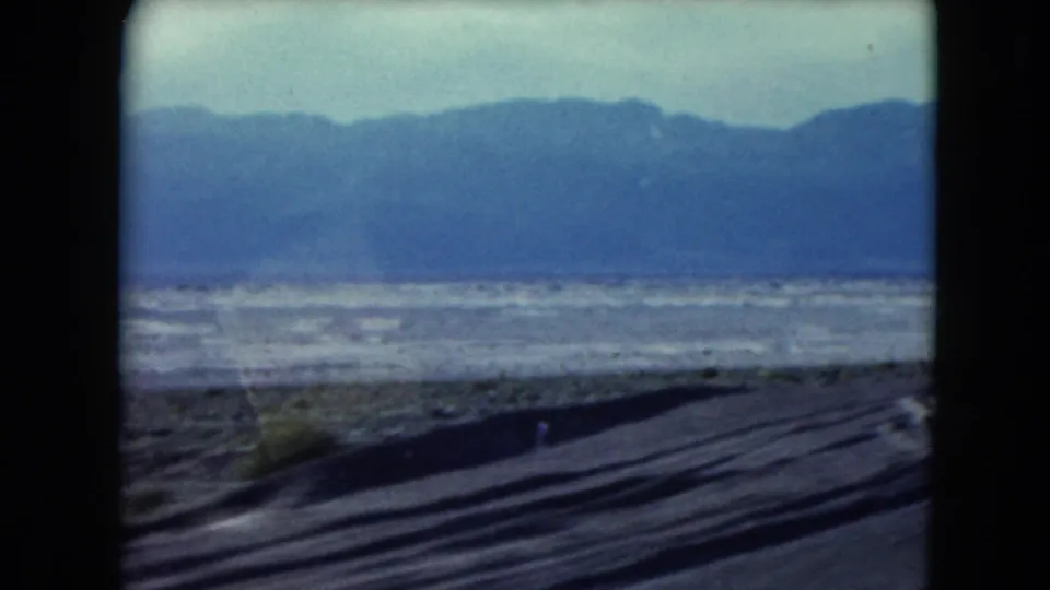1961 Nevada home-movie frame showing roadside desert, flat open ground, and distant mountains from a moving car.