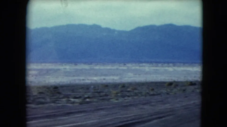 Wide 8mm view of Nevada desert terrain and a long blue mountain ridge, filmed from a passing vehicle in 1961.