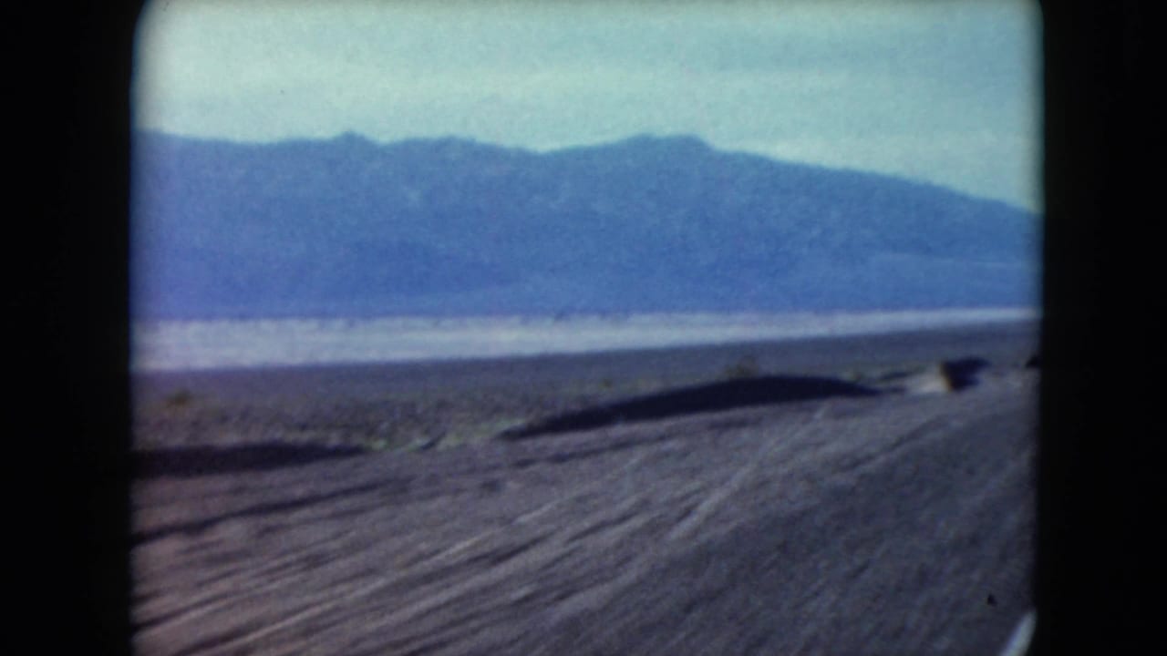 Desert highway shoulder with low scrub and mountains beyond the road.