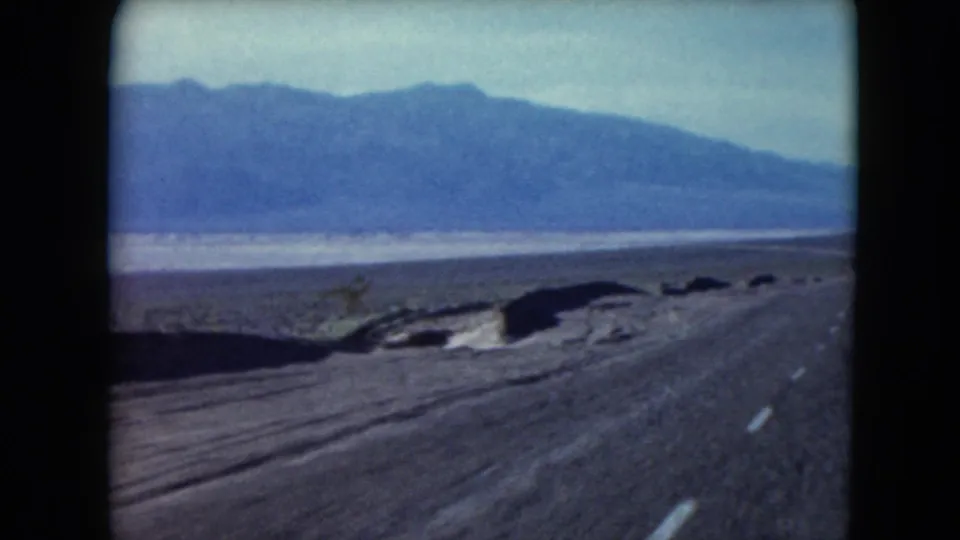 1961 Nevada home-movie frame showing a paved desert highway, rough shoulder, and mountain backdrop.