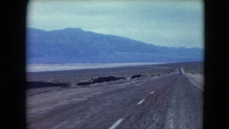 Nevada desert highway recedes toward mountains, with a lone distant vehicle visible ahead.