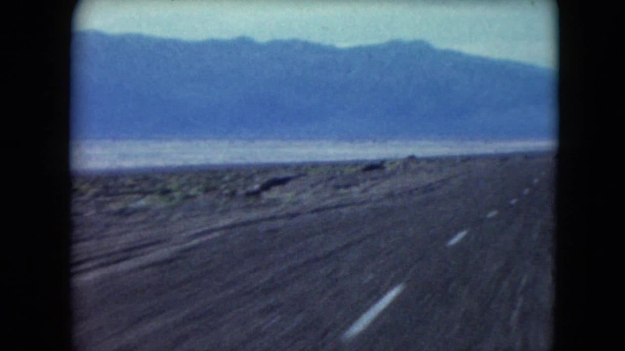 Desert highway receding toward blue mountains with sparse roadside vegetation.