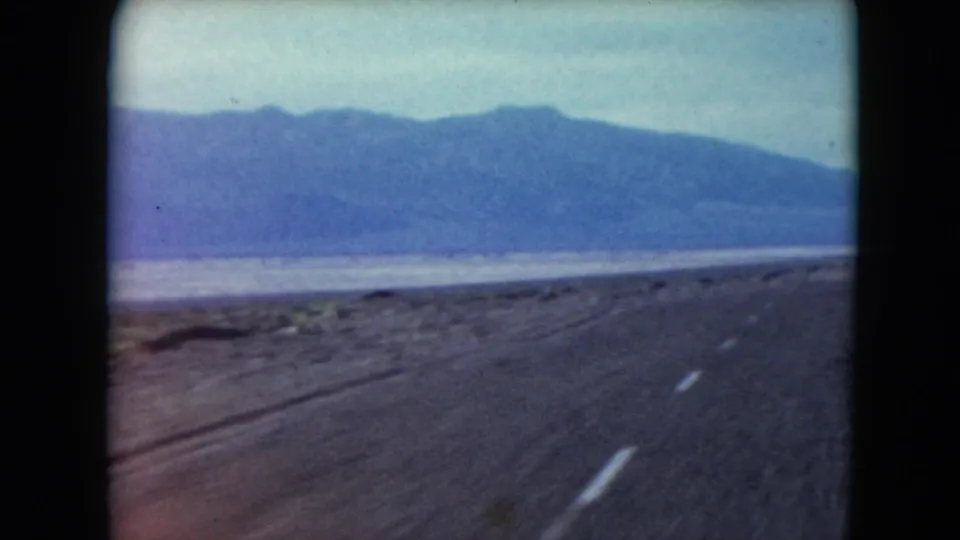 1961 Nevada 8mm footage frames the roadside and distant ridge from a vehicle traveling through desert terrain.