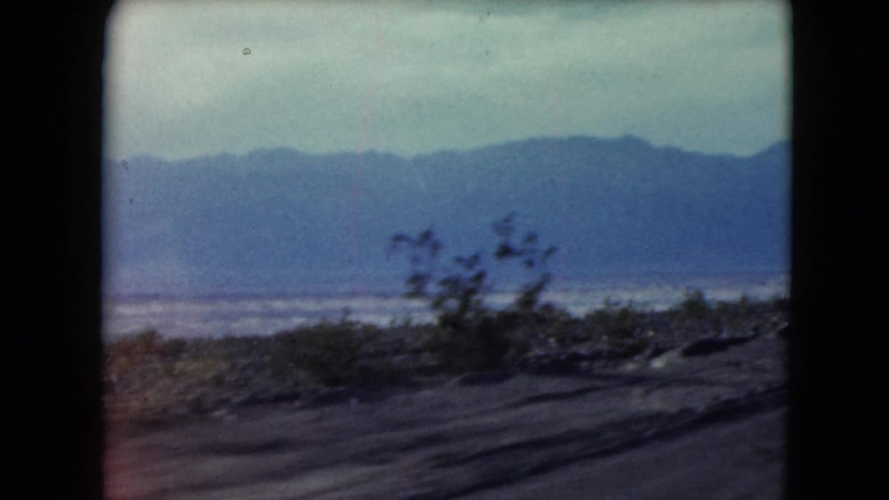 1961 Nevada 8mm frame showing a moving roadside view of desert shrubs, distant blue mountains, and a pale lake or salt flat.
