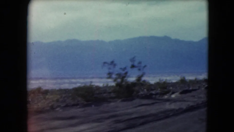 1961 Nevada 8mm frame showing a moving roadside view of desert shrubs, distant blue mountains, and a pale lake or salt flat.
