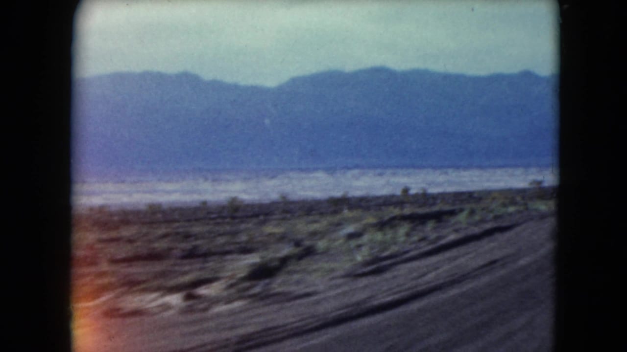 1961 Nevada roadside frame with a hazy mountain ridge, pale lake or salt-flat area, sparse scrub, and motion-blurred road edge.
