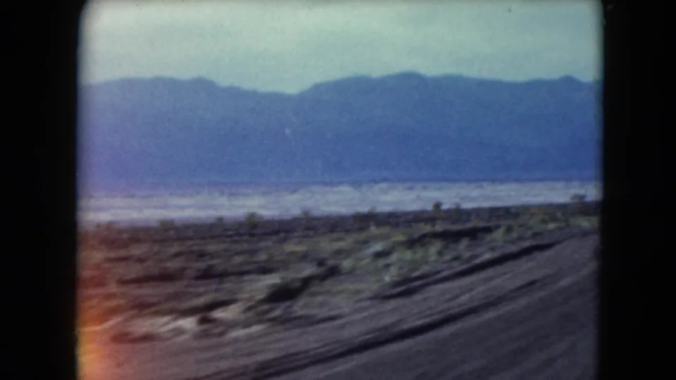 1961 Nevada roadside frame with a hazy mountain ridge, pale lake or salt-flat area, sparse scrub, and motion-blurred road edge.
