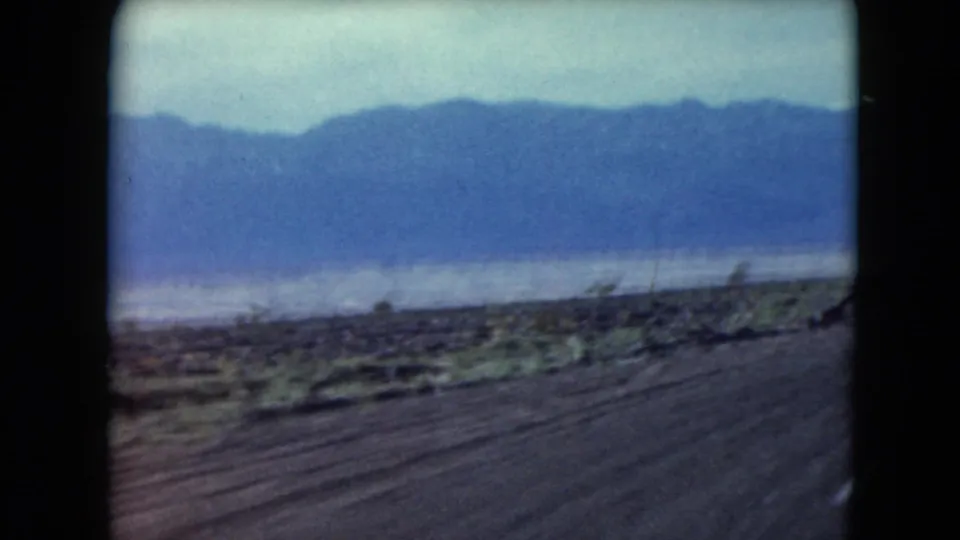 1961 Nevada 8mm view across a flat lake or salt-flat basin, with distant mountains and blurred roadside vegetation in front.