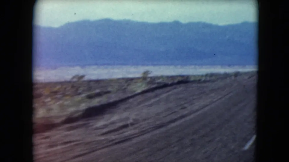 1961 Nevada home-movie frame showing a passing desert road edge, scattered scrub, a pale basin, and distant blue mountains.