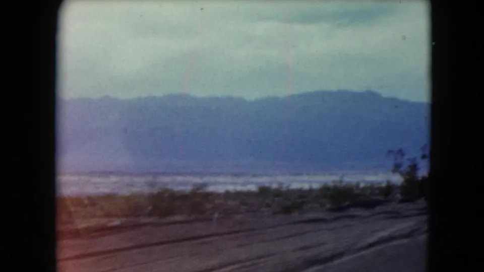 1961 Nevada 8mm frame showing a dirt desert road, sparse vegetation, and a distant mountain range under a hazy sky.