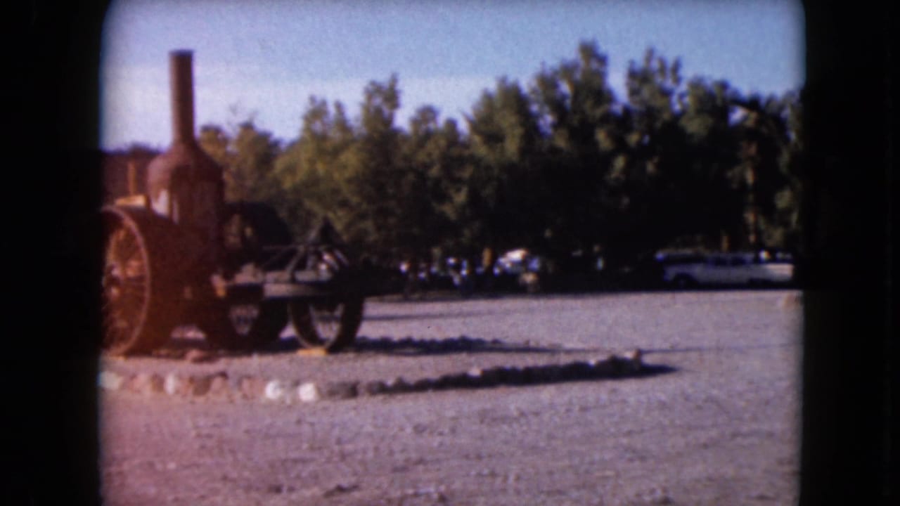 Steam traction engine displayed on a gravel lot with trees and parked cars behind it.