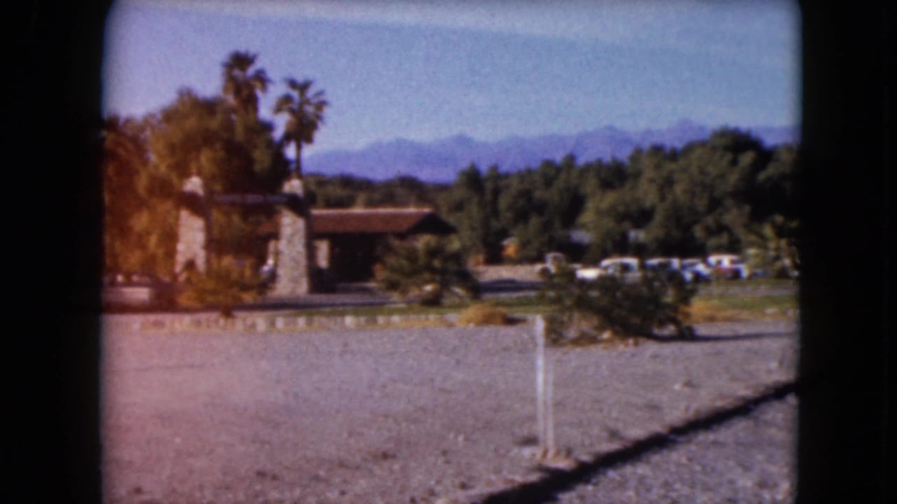 1961 desert resort entrance view with stone gateposts, palm trees, gravel parking, cars, and mountains beyond.