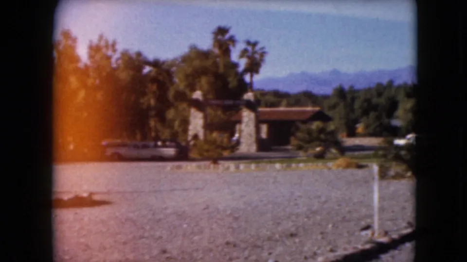 1961 8mm visitor view across a gravel foreground toward the Furnace Creek Ranch entrance, palms, car, and distant mountains.