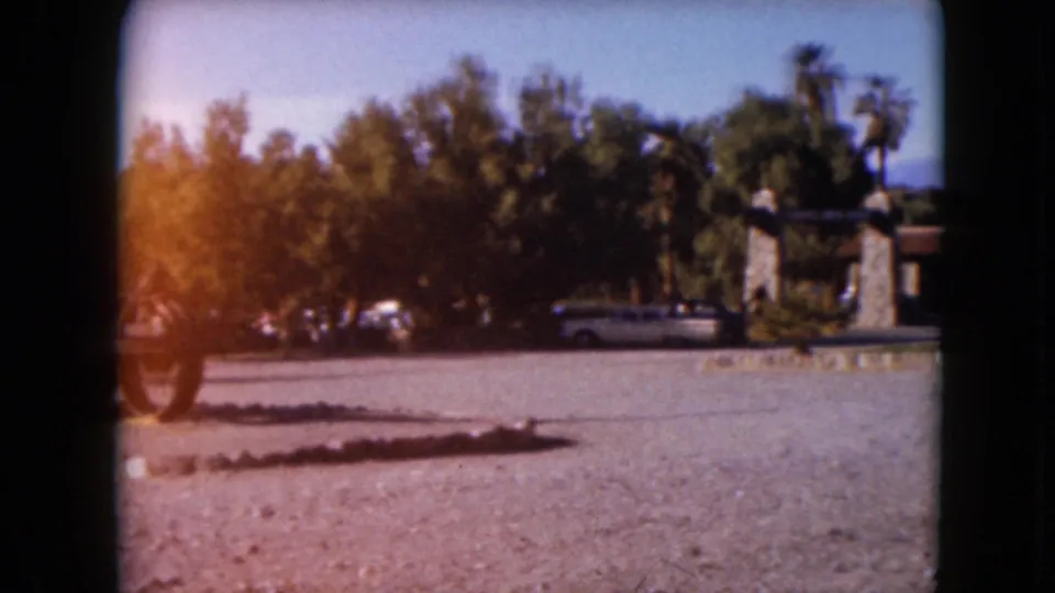 1961 8mm frame showing a wider Furnace Creek Ranch entrance view with gravel lot, trees, palms, parked car, and stone gateposts.