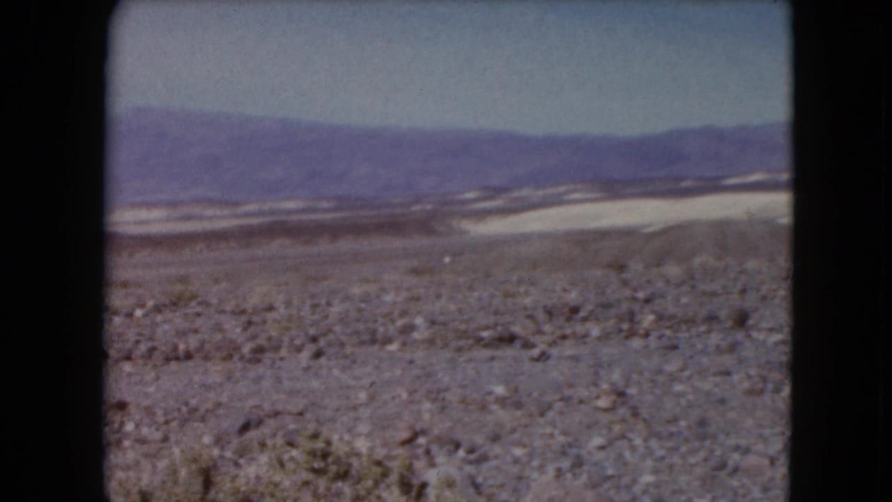 1961 8mm frame of rocky desert terrain stretching toward distant purple mountains under a pale sky in the Nevada travel sequence.