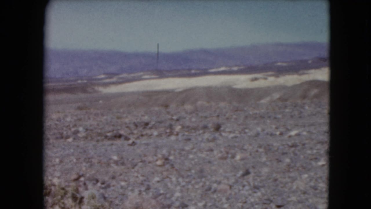 1961 8mm frame showing a desert valley view with rocky foreground, pale flat terrain, and distant mountains.