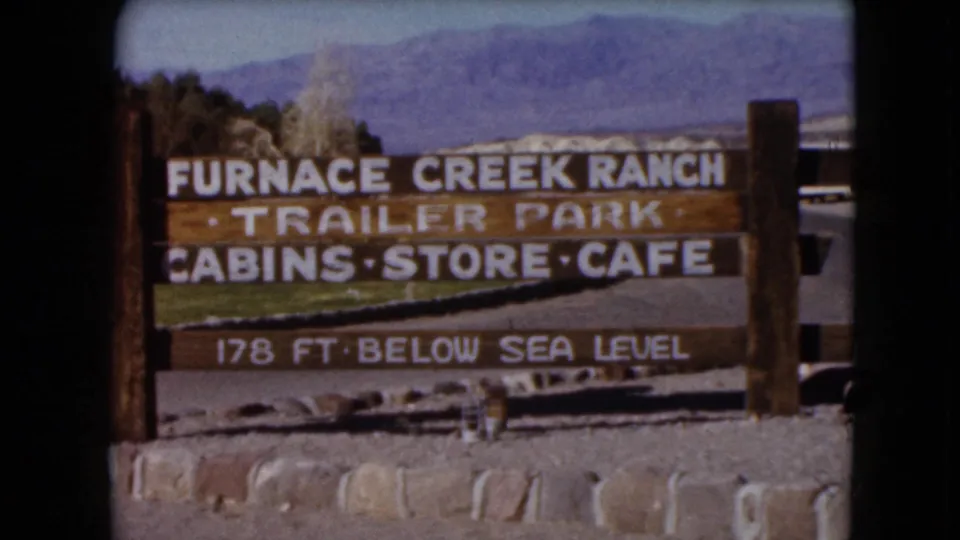 1961 8mm frame of a wooden Furnace Creek Ranch sign listing trailer park, cabins, store, cafe, and 178 ft below sea level.