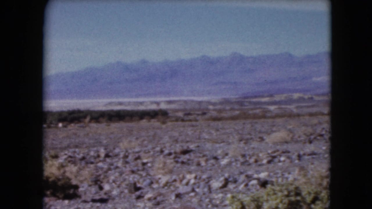 1961 Nevada home-movie frame showing rocky desert ground, scattered low vegetation, and distant purple mountain ridges.