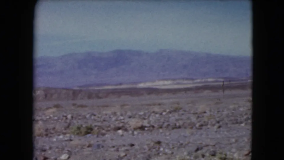 1961 Nevada landscape frame showing stony foreground, sparse vegetation, pale flats, and distant mountains.