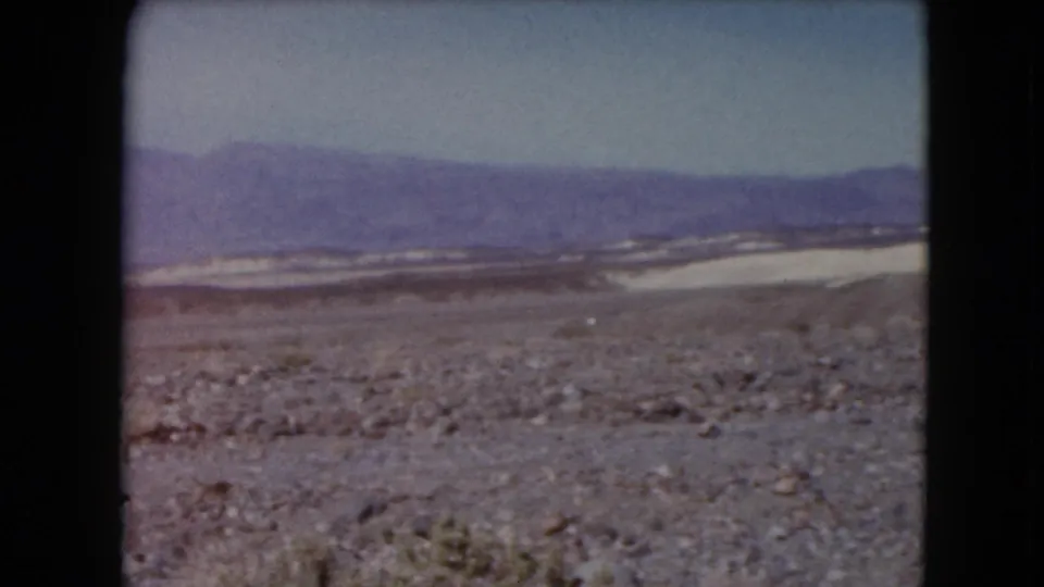 1961 Nevada home-movie frame with rocky desert basin, pale exposed flats, and purple-toned mountains on the horizon.
