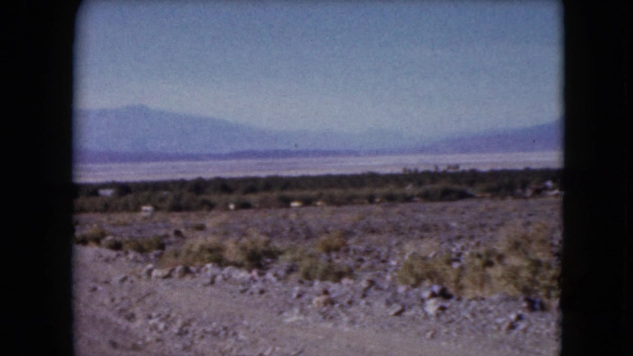 1961 Nevada 8mm frame showing rocky desert foreground, sparse shrubs, a pale distant flat, and hazy mountain ranges under blue sky.