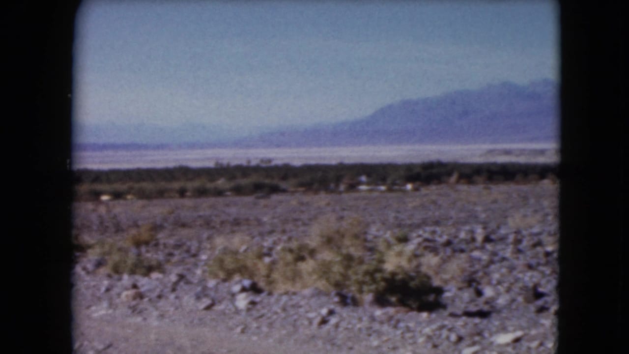 1961 Nevada home-movie frame with stony desert ground, scattered brush, a distant salt-flat-like band, and mountains beyond.