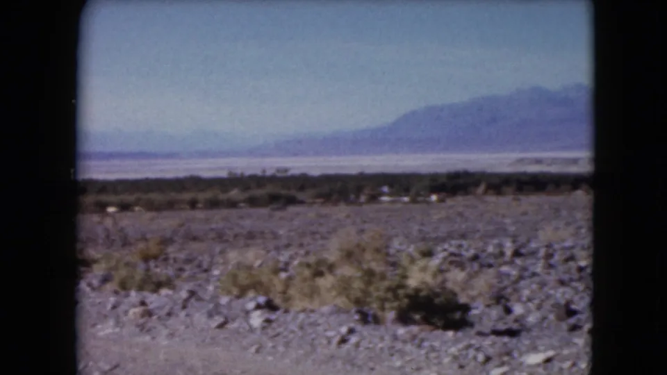1961 Nevada home-movie frame with stony desert ground, scattered brush, a distant salt-flat-like band, and mountains beyond.