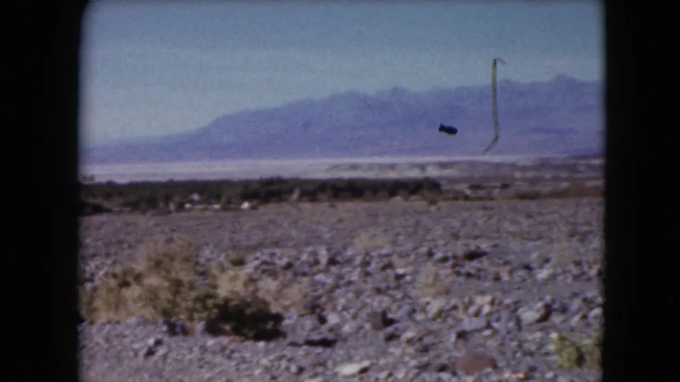 1961 Nevada 8mm frame showing a rocky desert slope, sparse brush, a distant light-toned flat, hazy mountains, and visible film marks.