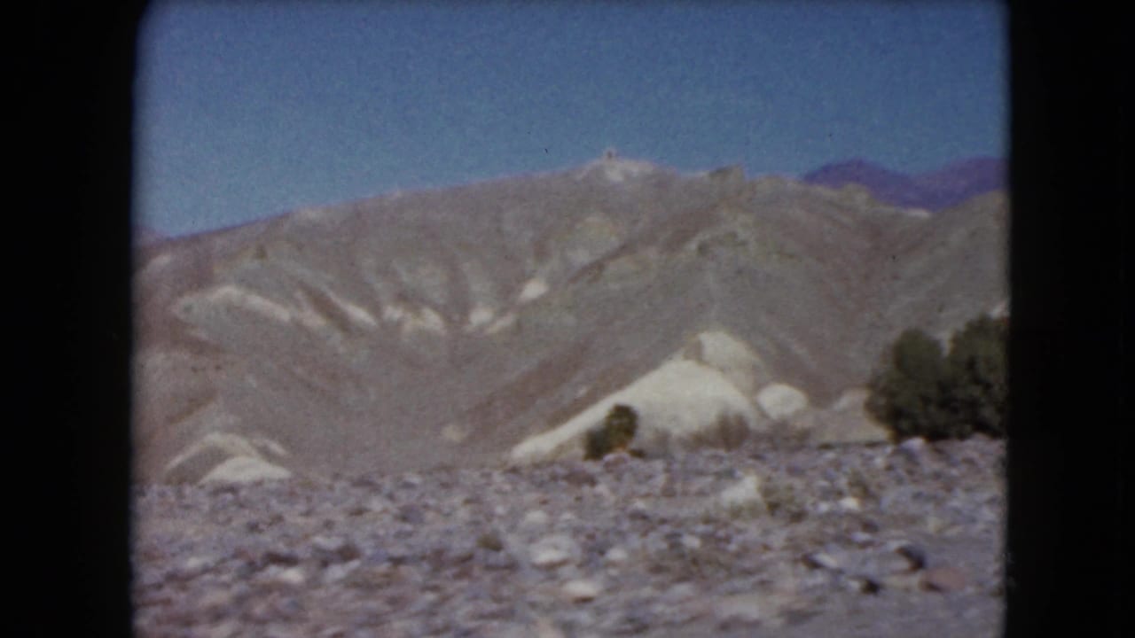 Rocky desert foreground with pale hills, distant mountains, shrubs, and blue sky.