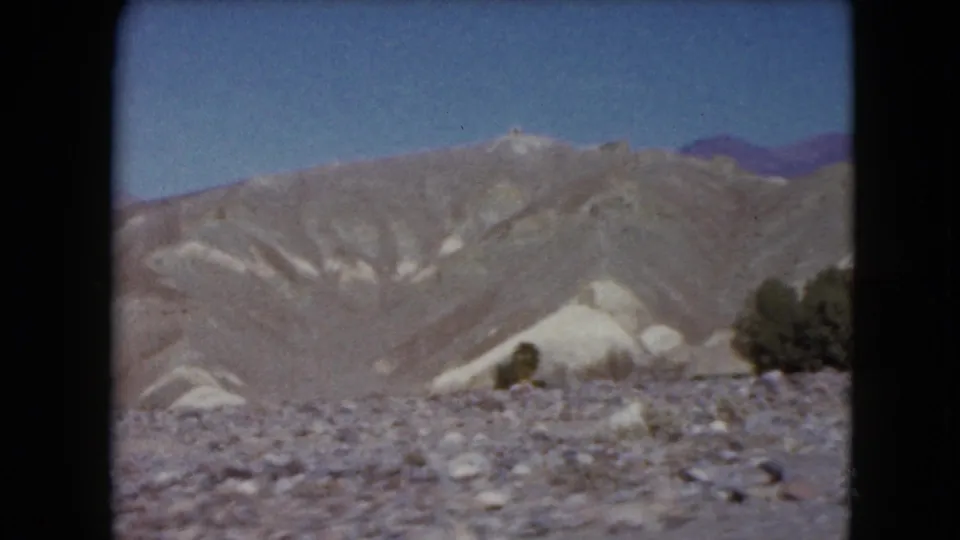 1961 Nevada 8mm frame showing rocky desert terrain, pale hills, sparse vegetation, and distant mountains under a clear sky.