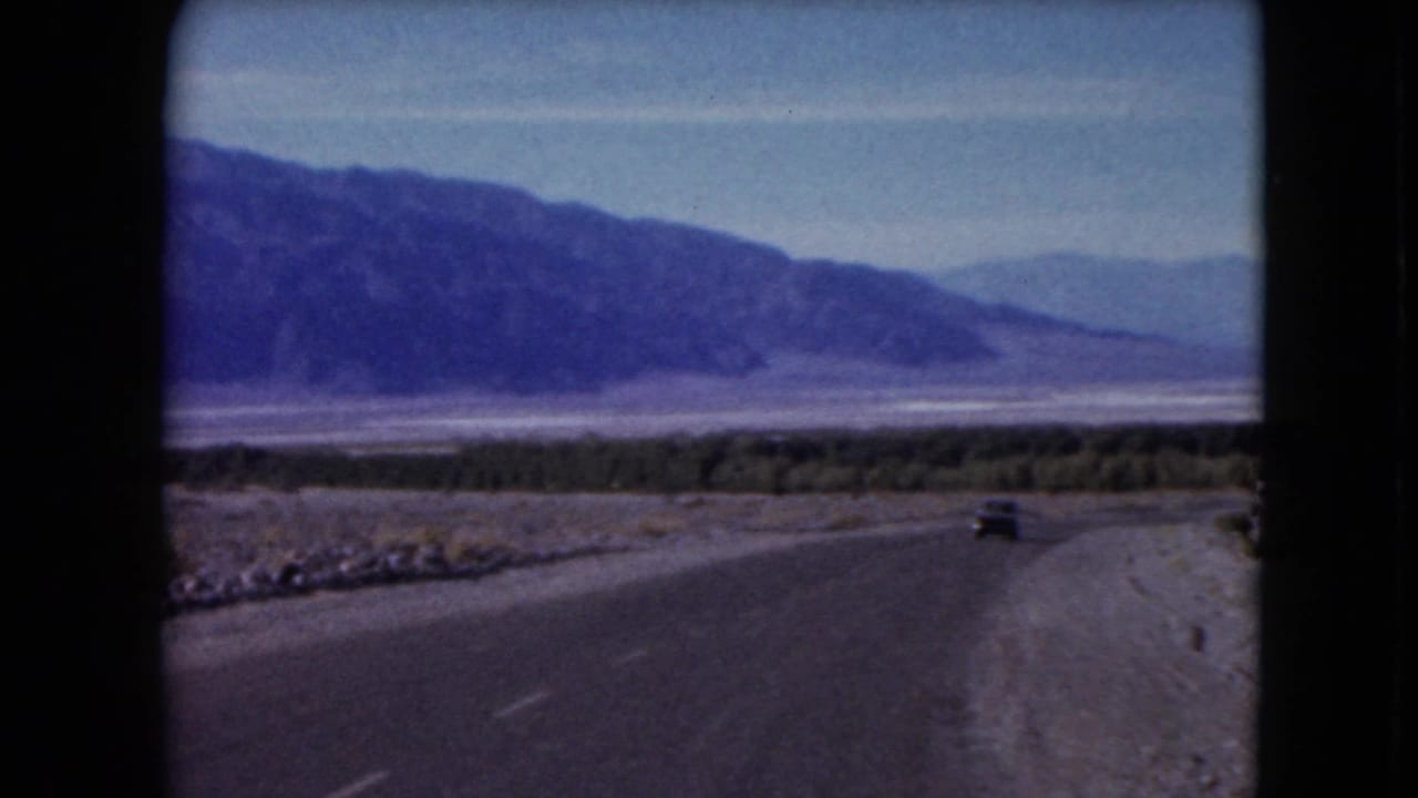 1961 Nevada road scene with a small car driving away along a paved desert road toward distant mountain ranges.