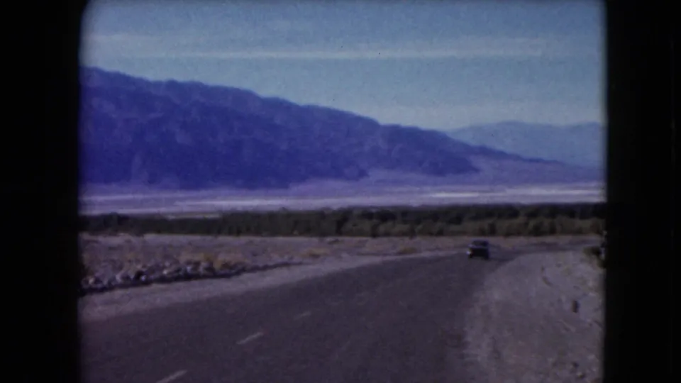 1961 Nevada road scene with a small car driving away along a paved desert road toward distant mountain ranges.