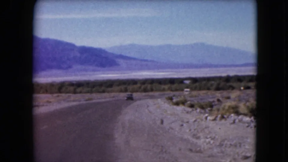 8mm home-movie view of a Nevada desert road curving through a valley, with a small car and layered mountains beyond.