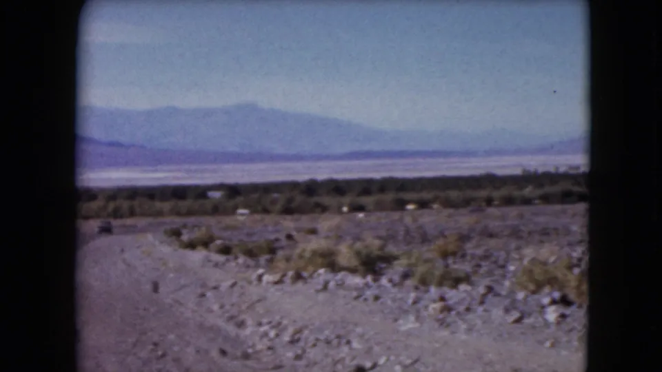 1961 Nevada desert landscape with rocky roadside ground, sparse brush, a valley floor, and hazy mountains in the distance.