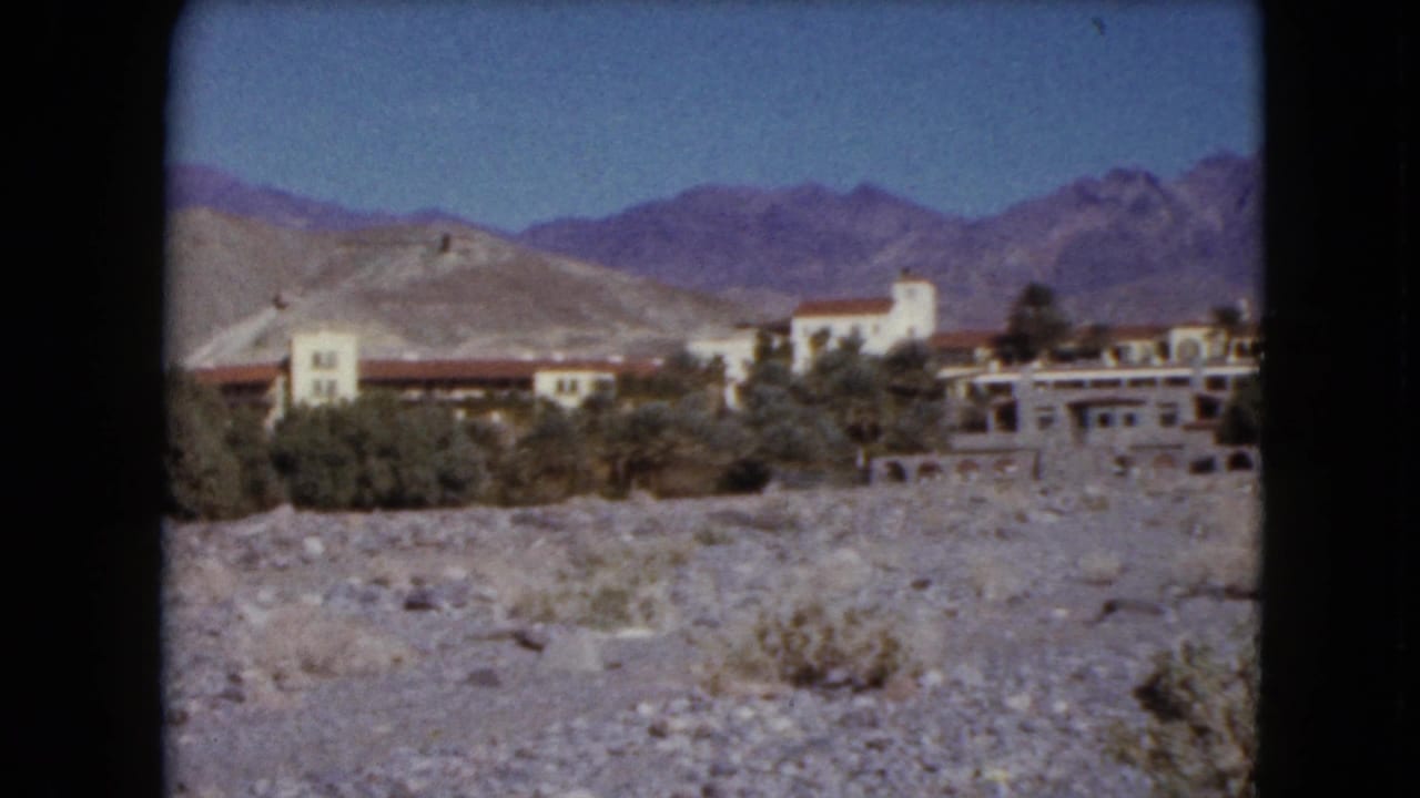 1961 Nevada 8mm frame showing red-roofed buildings, pale arid hills, distant mountains, blue sky, and gravelly desert ground.