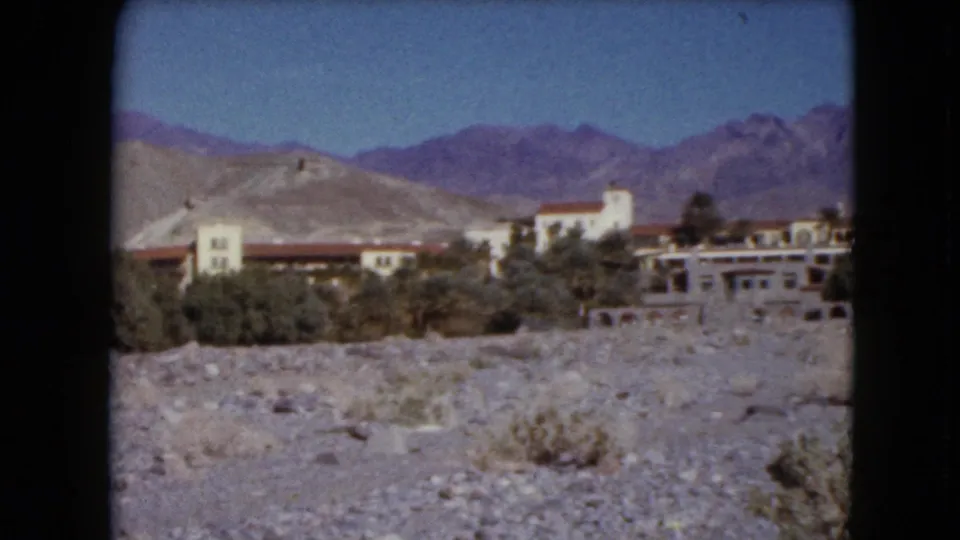 1961 Nevada 8mm frame showing red-roofed buildings, pale arid hills, distant mountains, blue sky, and gravelly desert ground.