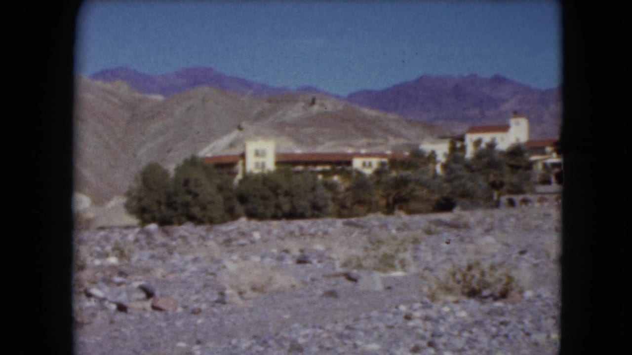 1961 Nevada home-movie view of a distant desert building complex framed by rocky ground, shrubs, pale hills, and purple-blue mountains.