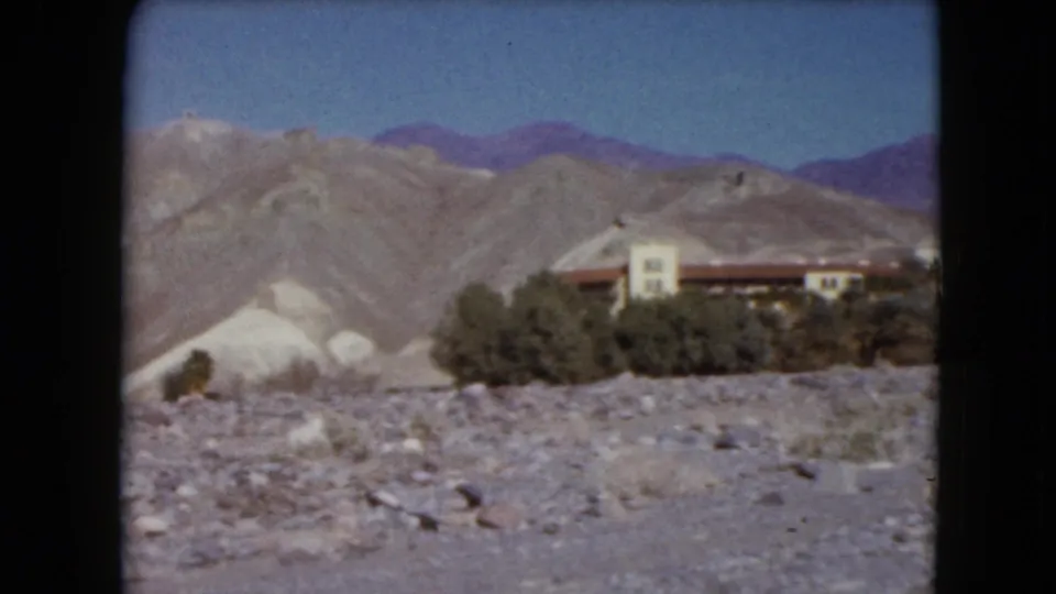 1961 Nevada 8mm frame with rocky foreground, scrub vegetation, a partial low building, pale hills, distant mountains, and clear blue sky.