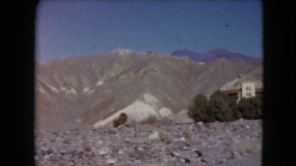 1961 Nevada desert view emphasizing pale hills, gravelly foreground, shrubs, blue sky, and the edge of a red-roofed building complex.