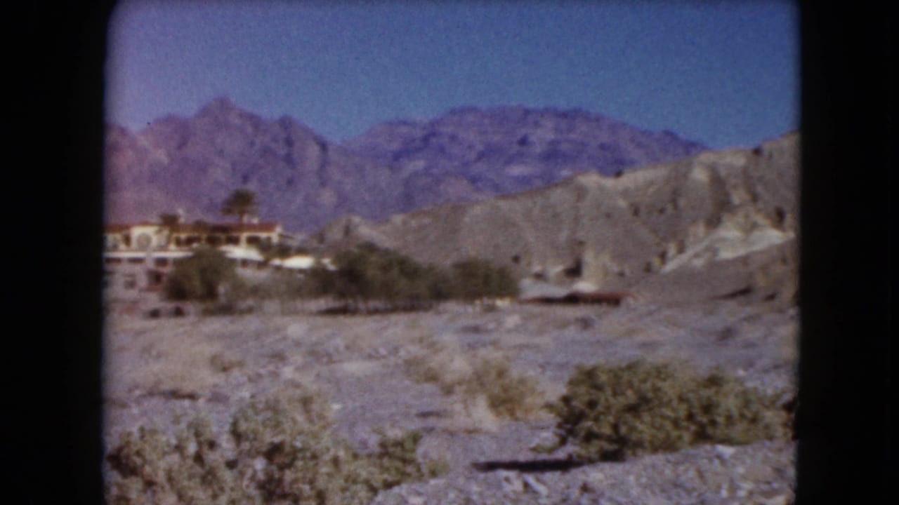 Distant resort buildings below purple mountains across a rocky desert foreground.