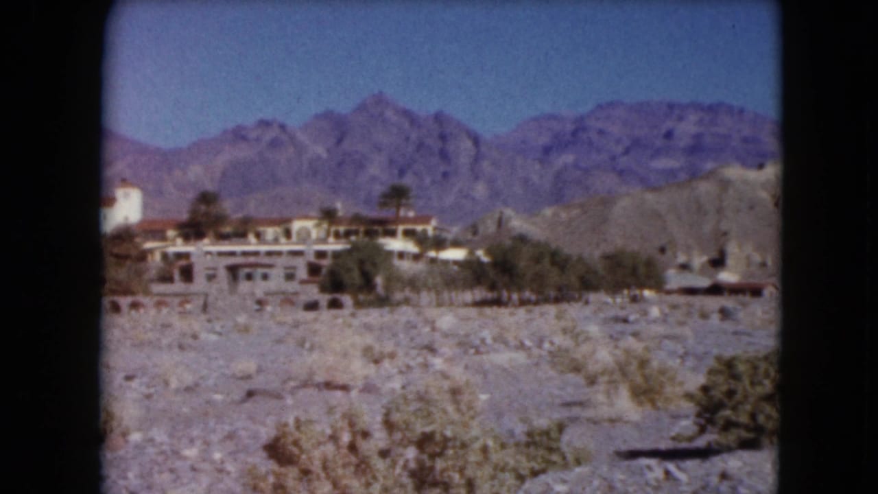 1961 Nevada 8mm view of a low resort complex with palms, pale walls, rocky desert terrain, and distant mountains.