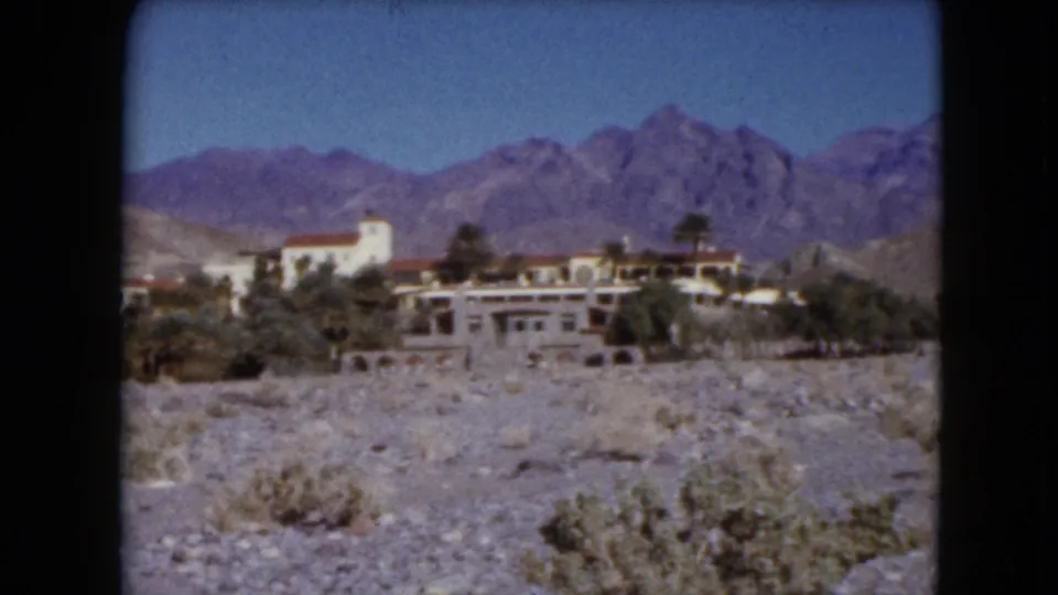 1961 Nevada home-movie frame with resort buildings, a pale tower, palms, scrub desert, and purple-toned mountains.