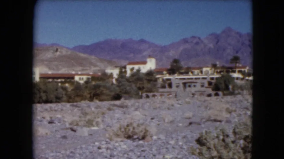 1961 Nevada 8mm frame showing resort-style buildings, scrub vegetation, stony desert terrain, and a broad mountain range.