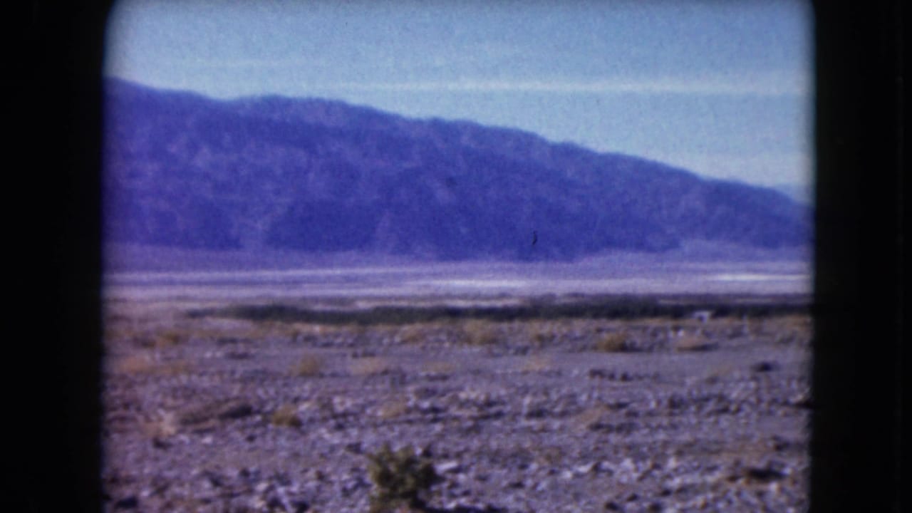 1961 Nevada 8mm frame showing a rocky desert foreground, pale basin, and dark distant mountain ridge beneath a clear sky.