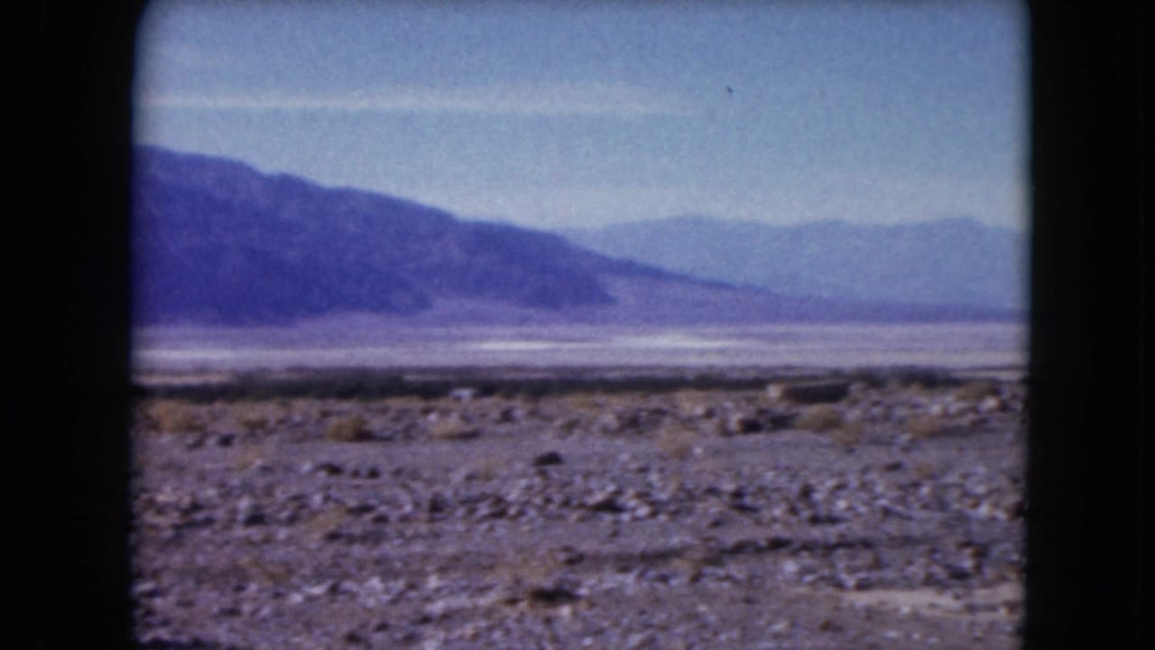 1961 Nevada desert view with scattered stones and brush in the foreground and hazy mountain ranges across the horizon.