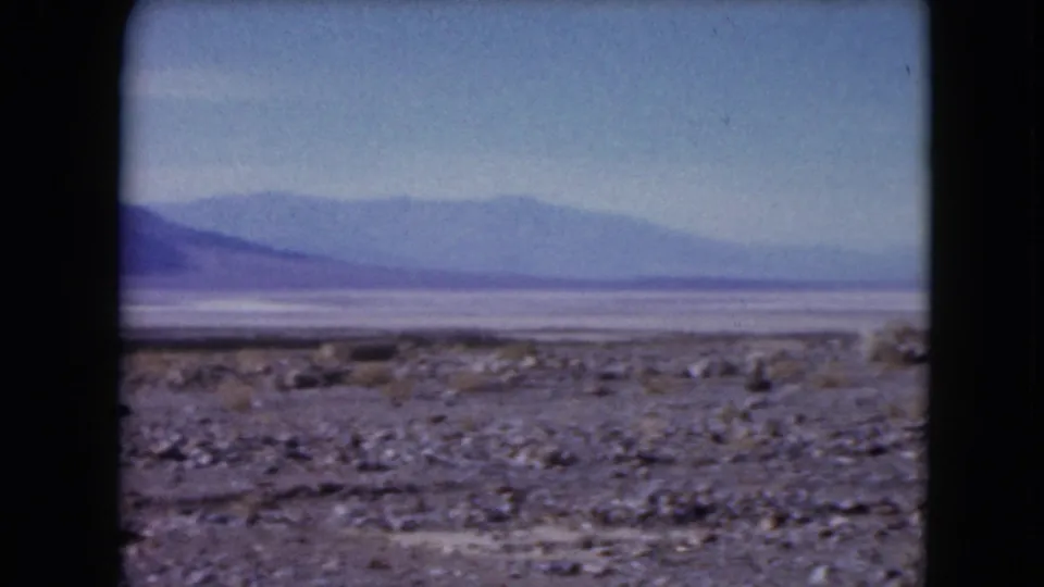 Soft 8mm frame of a Nevada desert basin, with rocky ground and scrub leading toward pale flats and distant blue mountains.